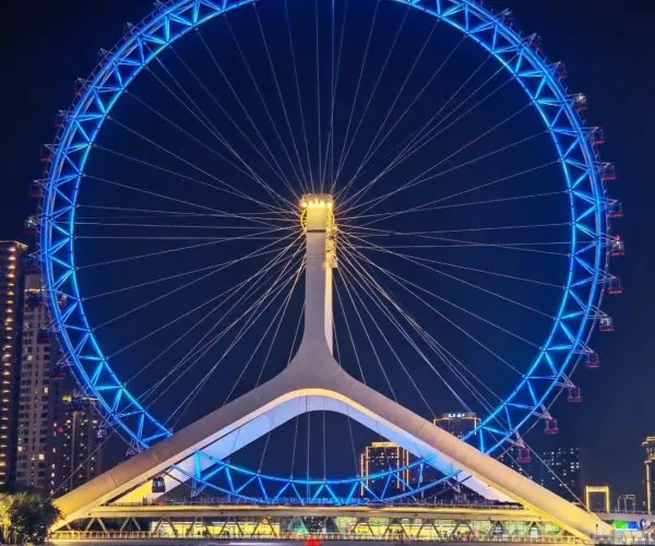 Nighttime view of a large blue-lit Ferris wheel over a river, with a white supporting pod and city lights in the background.