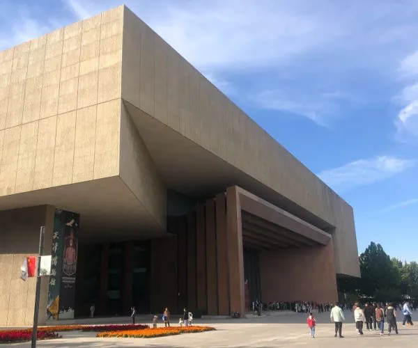 Large contemporary beige building with angular overhang, people walking in a wide plaza and a flower bed in the foreground.”