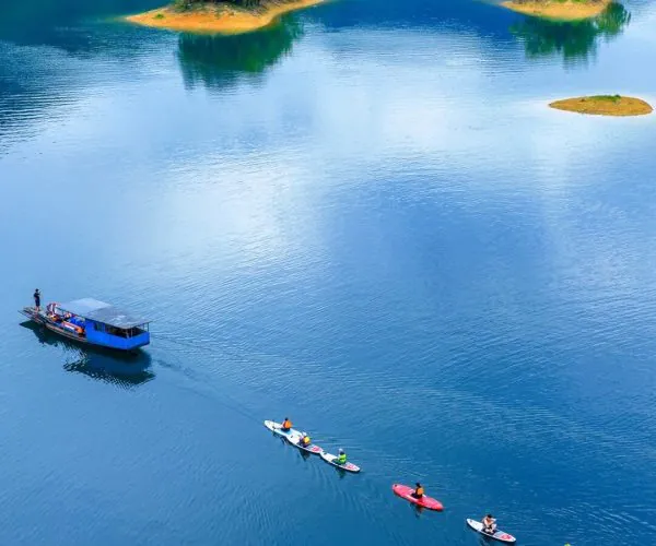 Taiping Lake,Huangshan,Chinatravel A calm blue lake with several small tree-covered islands and a blue boat on the left; people kayak in a line across the water.