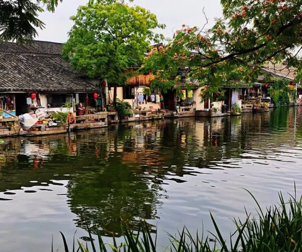 Anchang Ancient Town,Shaoxing,Chinatravel A canal lined with traditional houses, boats, and laundry drying on the shore, with green trees overhead and reflections in the water.