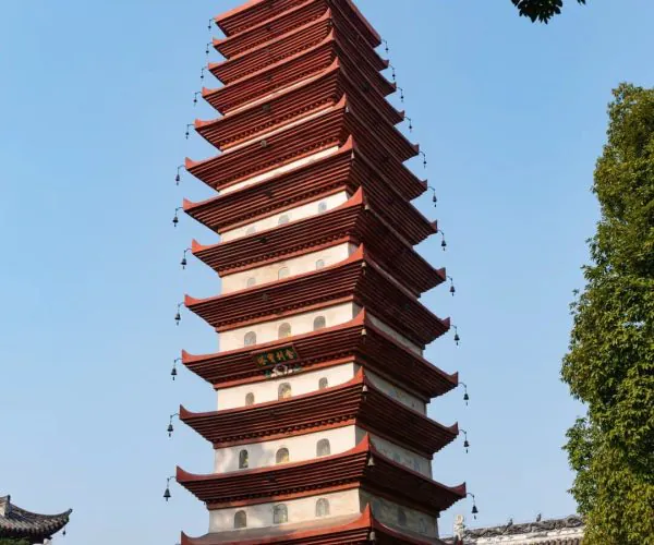 Baoguang Temple,Leshan,Chinatravel Tall multi-tiered Chinese pagoda with red eaves and hanging bells, set beside temple buildings under a blue sky.