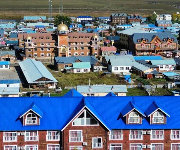 Shiwei Russian Ethnic Township,Hulunbuir,Chinatrip Aerial view of a town with bright blue roofs and brick buildings, with mountains in the distance.