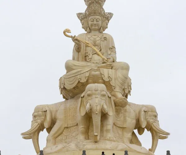 Mount Emei Scenic Area,Leshan,Chinatravel Large beige statue of a multi-headed deity seated on an elephant throne, atop a stepped platform, holding a curved staff against a pale sky.