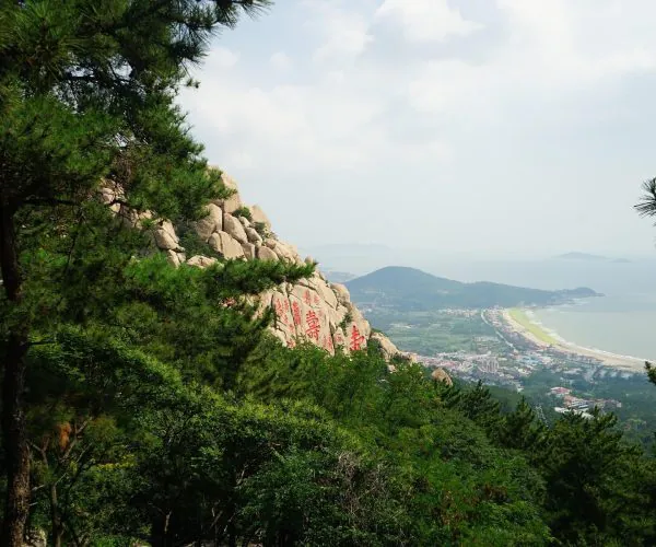 Mount Laoshan,Qingdao,Chinatravel Coastal town below a rocky hillside with boulders carved with red Chinese characters, viewed through pine trees.