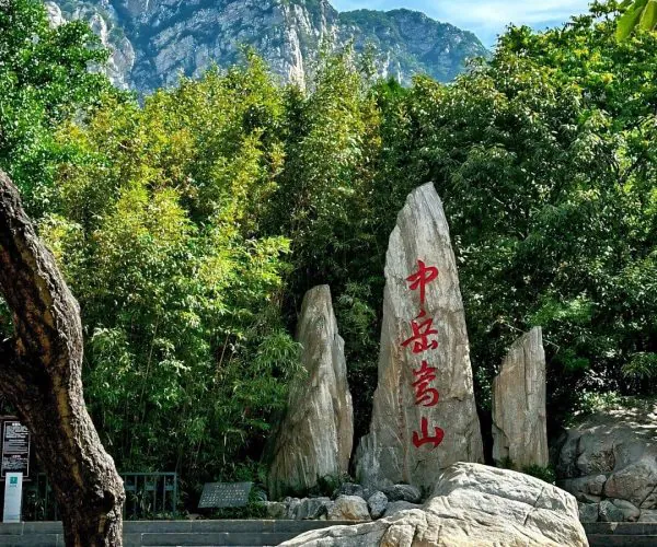 Songshan (Song Mountains) Scenic Area,Shaolin & Songshan Area,Chinatrip Mountain landscape with tall rocky peak behind lush green bushes and a vertical rock slab with red Chinese characters; a rocky foreground and steps hint a park entrance