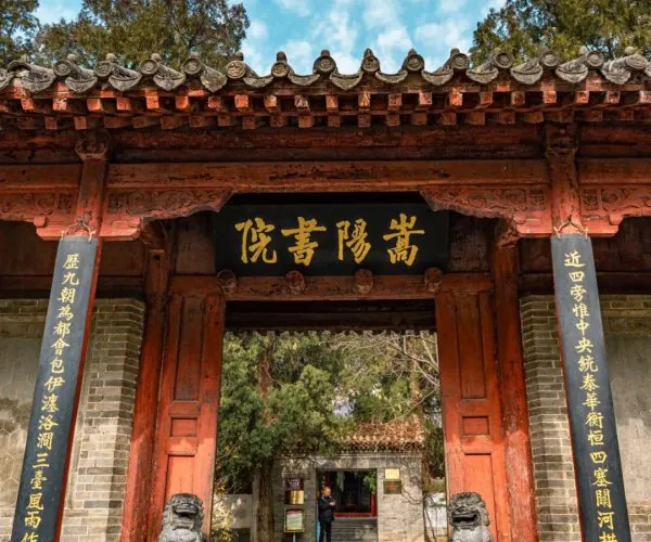 Songyang Academy,Shaolin & Songshan Area,Chinatrip Traditional Chinese arched gate with a red wooden frame, gold inscription plaque, and stone guardian lions flanking the stairs under a blue sky.