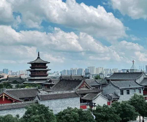 Pingcheng Ancient Town,Datong,Chinatravel Traditional Chinese temple complex with a multi-tier pagoda against a blue sky, row of grey-tiled roofs, and urban skyline in the distance behind green trees.
