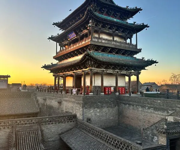 Pingyao Weather,Pingyao,Chinatravel Three-tier Chinese pagoda atop a brick fortress at sunset, with visitors along the walkway and decorative eaves visible in the sky-blue light.