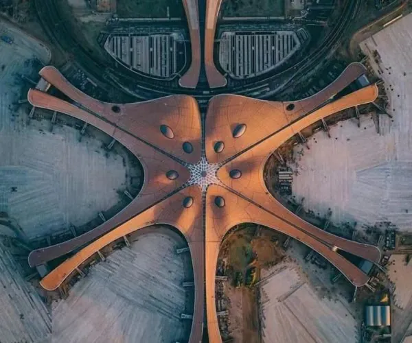 Beijing Daxing Airport, beijing, china, chinatrip, Aerial view of a starfish-shaped orange-roofed building with five long curved arms radiating from a central hub, set among a construction site and surrounding streets.
