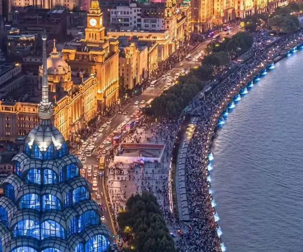 Aerial night view of Shanghai's Bund with illuminated historic buildings, a crowded riverside promenade, and a blue glass-domed building in the foreground.