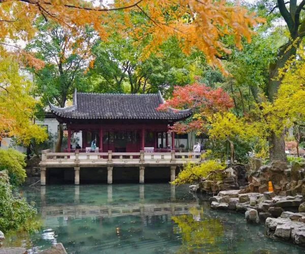 Traditional Chinese pavilion on stilts over a calm pond, surrounded by orange and green autumn leaves and trees.