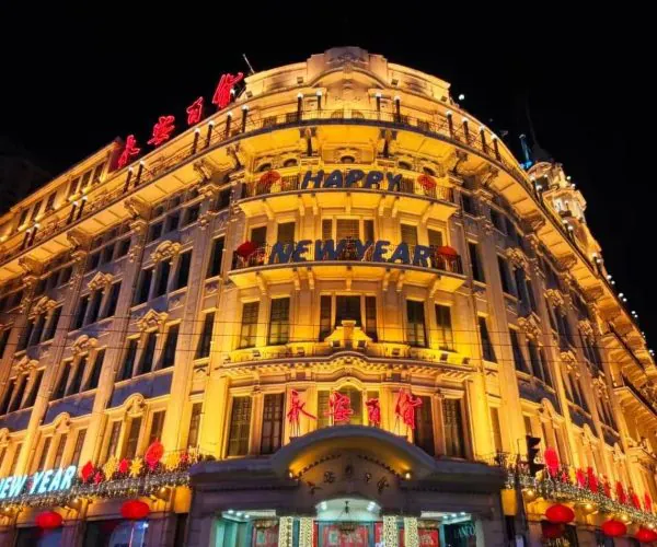 Ornate night-time building facade lit in warm yellow lights with 'Happy New Year' banners and red lanterns outside.
