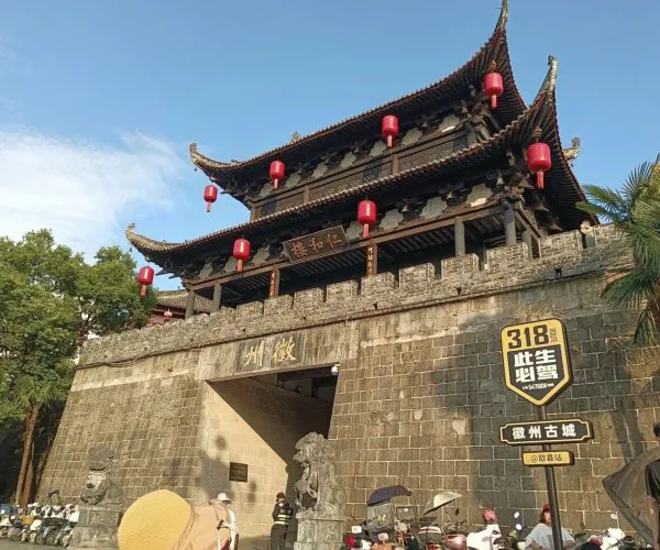 Huizhou Ancient City,Huangshan,Chinatravel Traditional Chinese gate with a multi-tiered roof and red lanterns hanging above a stone city wall, with people and scooters in the foreground.
