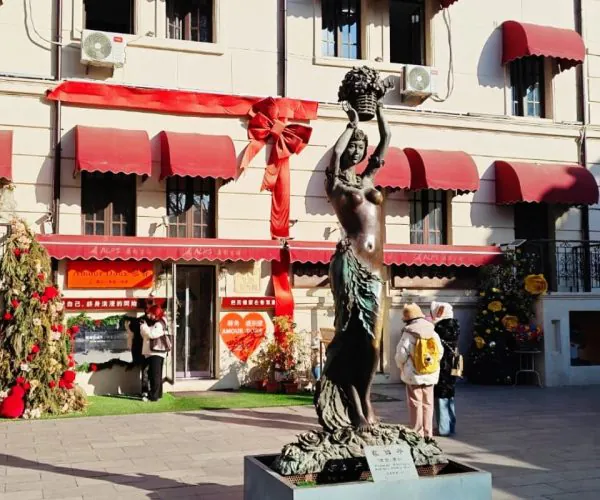 Bronze statue of a woman holding a bouquet atop a pedestal in a plaza, with a red-ribbon storefront behind.