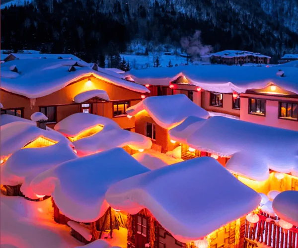 Enchanting Winter Wonderland: 3-Day Journey Through Harbin: China's Snow Kingdom, chinatrip, Snowy mountain village at night with warm orange lights glowing from windows and heavy snow on roofs under a starry blue sky.