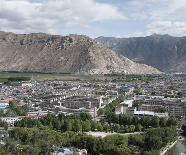 Lhasa Weather,Lhasa,Chinatravel Townscape at the foot of rugged mountains; dense low-rise buildings, tree-lined streets, and a river/park in the foreground.