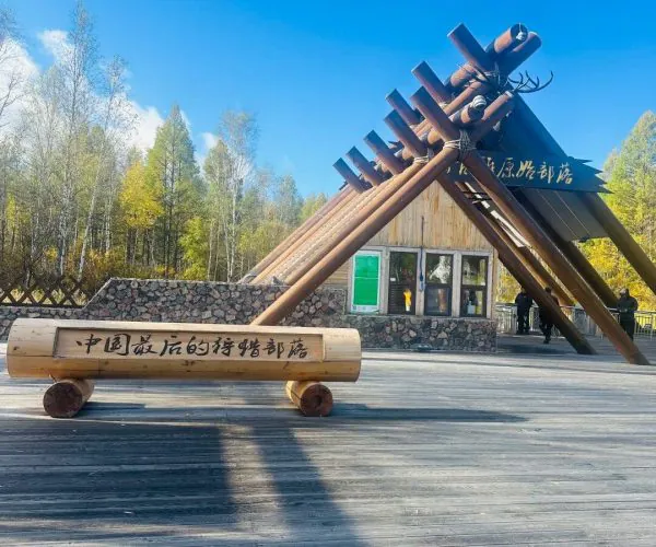 Aoluguya Reindeer Tribe,Hulunbuir,Chinatrip Outdoor wooden sculpture on a plaza: a triangular arch formed by thick logs, with a bench inscribed in Chinese and a small building in the background among trees on a sunny day.