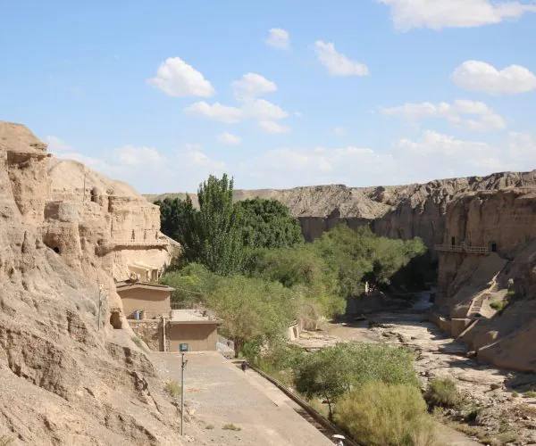 Dunhuang Weather,Dunhuang,Chinatrip Desert canyon with beige cliffside buildings carved into rock, a dry riverbed, and green trees along a path