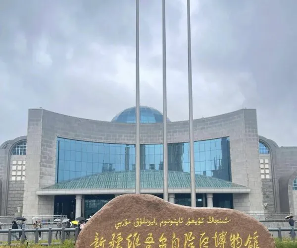 Xinjiang Regional Museum,Urumqi,Chinatrip Stone with gold inscription in Chinese and Uyghur in front of a modern museum building with a glass facade and a blue-green dome, flanked by flagpoles under a cloudy sky.