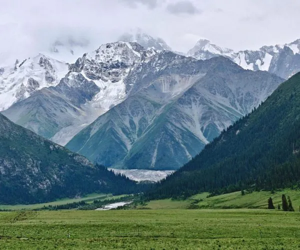 Xinjiang Weather,Xinjiang,China tour Snow-capped mountains rise above a green valley with a meadow in the foreground.