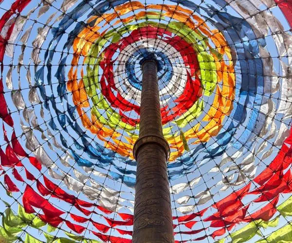 Riyue Mountain Pass,Xining,Chinatrip Tall central pillar surrounded by concentric rings of colorful prayer flags in red, orange, yellow, green, blue, and white, against a blue sky.
