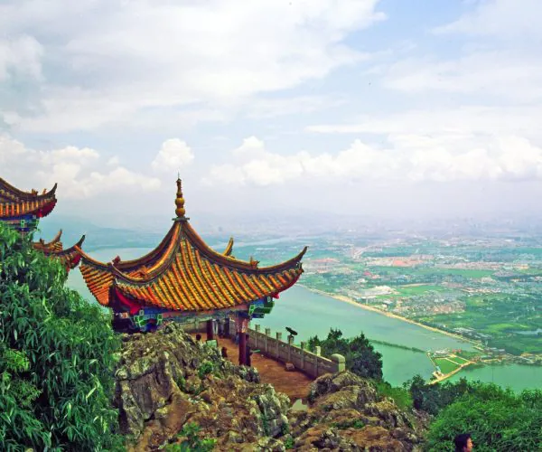 Kunming Weather,Kunming, Chinatravel Traditional Chinese temple with orange tiled curved roofs on a rocky hilltop overlooking a river and distant cityscape.