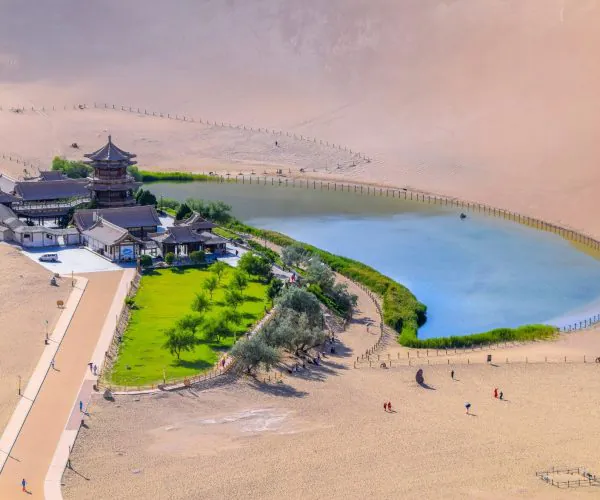Crescent Lake & Mingsha Mountain,Dunhuang,Chinatrip Aerial view of a pagoda-style temple complex beside a green lawn and calm blue lake, with vast sandy dunes in the background.