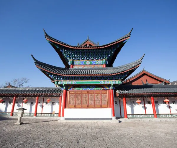 Mu’s Residence,Lijiang,Chinatravel Traditional Chinese temple gate with layered, upturned eaves, vibrant blue and green ornamentation, red pillars, and surrounding white walls under a clear blue sky