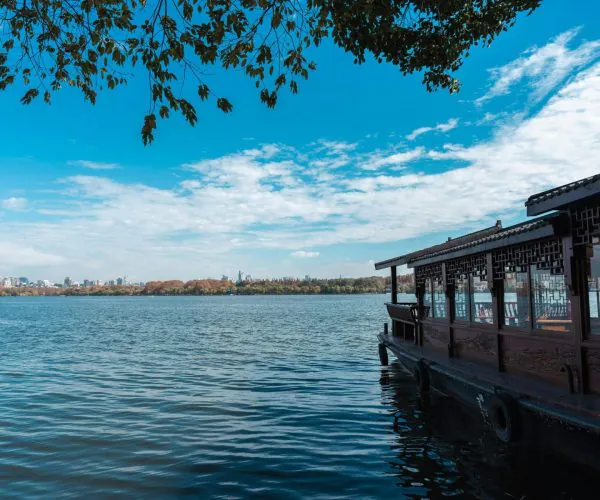Hangzhou,Zhejiang,Chinatravel Lakeside scene with a traditional wooden pavilion along the water, under tree branches and a blue sky with distant city skyline.