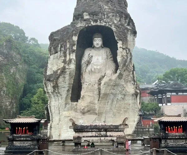 Keyan Scenic Area,Shaoxing,Chinatravel Large Buddha statue carved into a rock alcove within a cave, with temple buildings nearby on a rainy day.