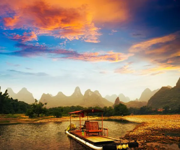 Guilin Weather,Guilin,Chinatravel Wooden raft with a red canopy on a calm river at sunset, karst mountains in the distance.