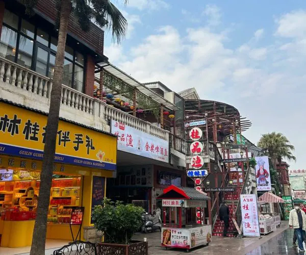 Zhengyang Pedestrian Street,Guilin,Chinatravel Busy shopping street with Chinese storefronts, a yellow shop front on the left, palm trees, and outdoor stalls under a blue sky.