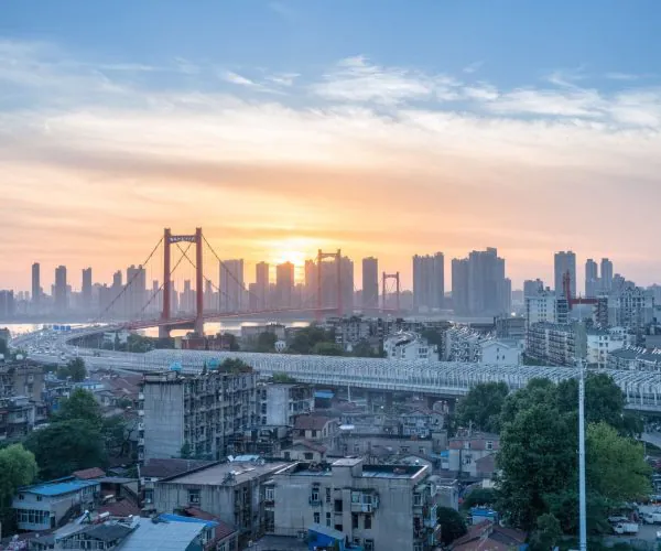 Wuhan Weather,Wuhan,Chinatravel Sunset cityscape with a red cable-stayed bridge over a river and a skyline of tall buildings, foreground filled with low-rise homes and trees.