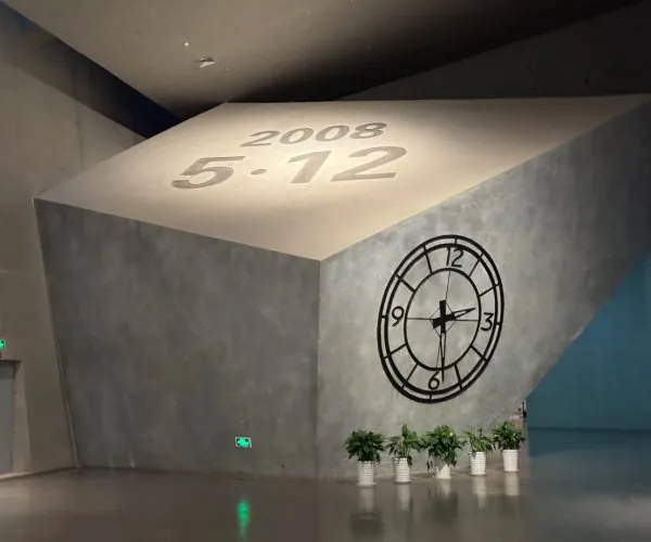 Jiangyou Earthquake Museum,Mianyang,Chinatravel Interior sculpture: a tilted concrete cube with '2008 5.12' inscribed on its top and a large clock graphic on the side, plants at the base.