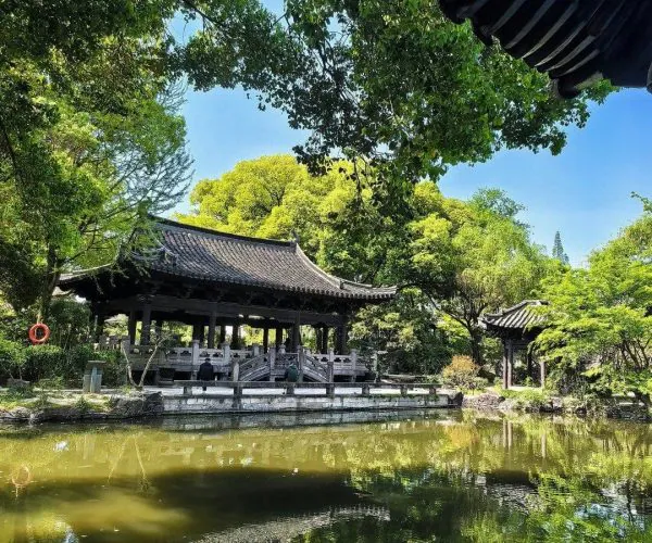 Shen Garden,Shaoxing,Chinatravel Traditional Chinese wooden pavilion by a calm pond, surrounded by lush green trees and a clear blue sky, with a stone walkway and reflections in the water.