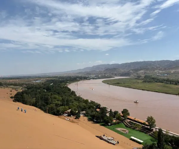 Shapotou Desert Scenic Area,Yinchuan,Chinatrip Panoramic view of a desert dune dropping to a brown river, with green vegetation, resort lawn, and distant mountains under a blue sky.