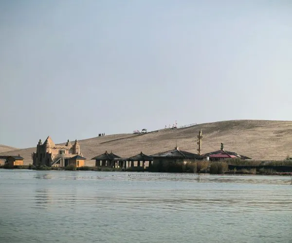 Shahu Lake (Sand Lake) Scenic Area,Yinchuan,Chinatrip Desert lakeside with a castle-like building and thatched-roof pavilions along calm water, and a sandy dune in the background.