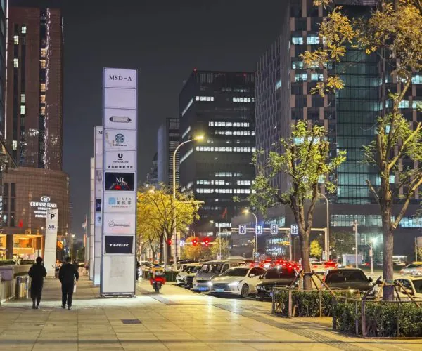 Night city street with illuminated buildings, parked cars, and pedestrians along a sidewalk with a tall signpost in the foreground.