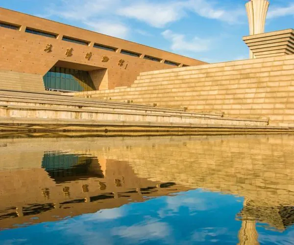 Luoyang Museum,Luoyang,Chinatravel Beige stone building with a tall sculptural column on the right, terraced steps, blue sky, and a reflecting pool showing the structure.",