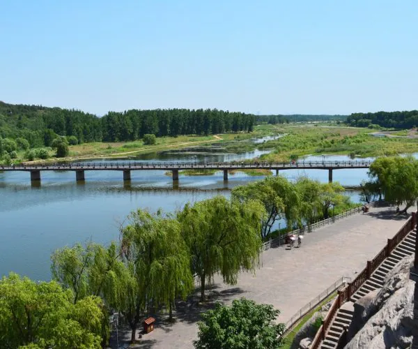 Luoyang Weather,Luoyang,Chinatravel Scenic riverside park with a long pedestrian bridge crossing calm water and green trees along the bank.