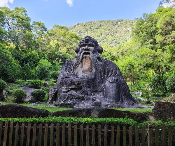Mount Qingyuan Scenic Area,Quanzhou,Chinatravel Large stone statue of a bearded sage seated in a garden, with trees and a wooden fence in the foreground.