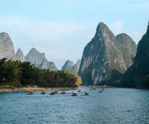 Li River,Guilin,Chinatravel Calm river lined with forested shore and groups of small boats, set against towering limestone mountains under a blue sky.
