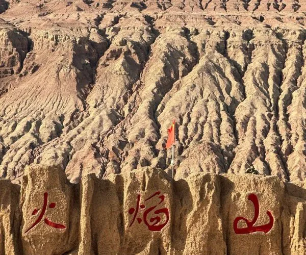 Flaming Mountains,Turpan,China tour Rugged desert landscape with jagged, eroded hills; in the foreground a textured stone barrier bearing three red Chinese characters and a small red flag.