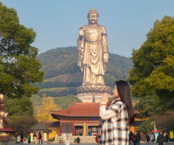 Lingshan Grand Buddha,Suzhou,Chinatravel Young woman in a plaid coat praying with pressed palms in front of a towering Buddha statue in a temple courtyard, trees in the background.