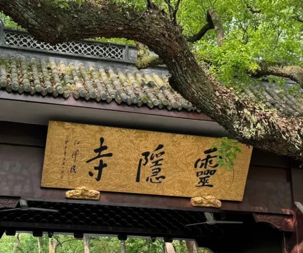 Lingyin Temple,Hangzhou,Chinatravel Golden plaque with black Chinese calligraphy mounted on a dark wooden gate, under a tiled roof, with a large tree branch arching overhead.