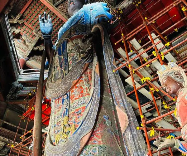 Large, intricately painted standing bodhisattva statue beneath red scaffolding in a temple, raised right hand signaling blessing.