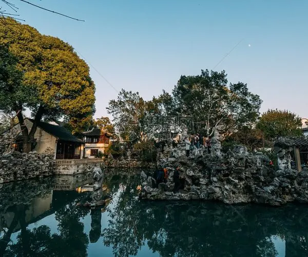 Lion Grove Garden,Suzhou,Chinatravel Visitors explore jagged rock formations around a reflective pond in a traditional Chinese garden, with a pavilion to the left and trees in the background.
