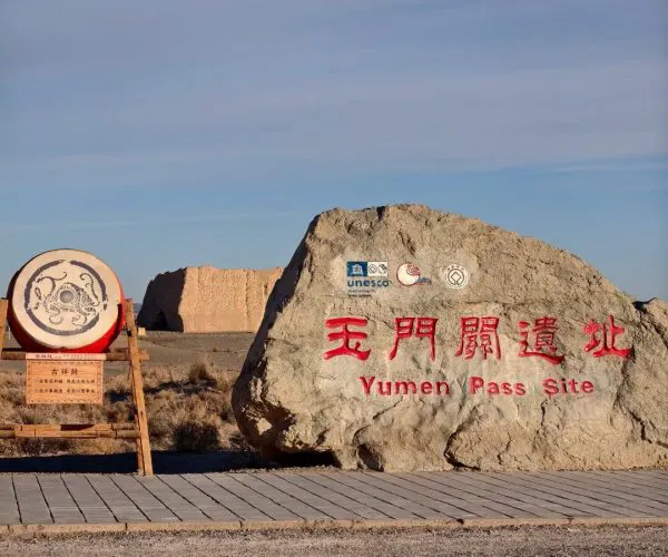 Yumen Pass,Dunhuang,Chinatrip Large rock with red Chinese characters and the words 'Yumen Pass Site' in English, with logos above, beside a wooden drum in a desert landscape.