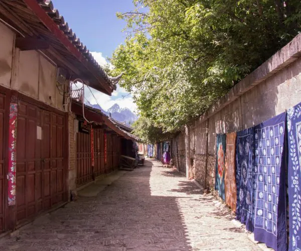 Baisha Murals & Baisha Village,Lijiang,Chinatravel Narrow village alley with wooden storefronts on the left and colorful fabrics hanging along a stone wall on the right, mountains in the distance.