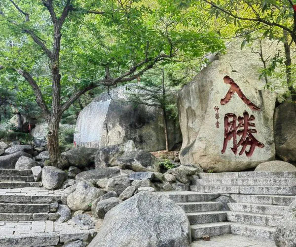 Stone steps lead up through a rocky garden with large boulders and a rock featuring red Chinese characters amid green trees.